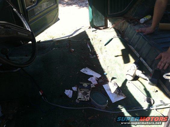 Interior of a vehicle with the carpet removed, showing the floor and door panel. Tools and parts are scattered around