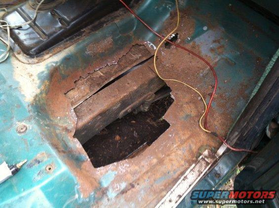Interior view of a rusted floor pan with a large hole exposing the undercarriage. Wires are visible