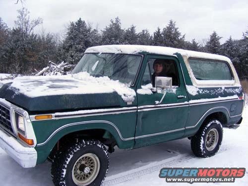 Green 1978 Ford Bronco with snow on hood and roof, person in driver's seat, snowy trees in background