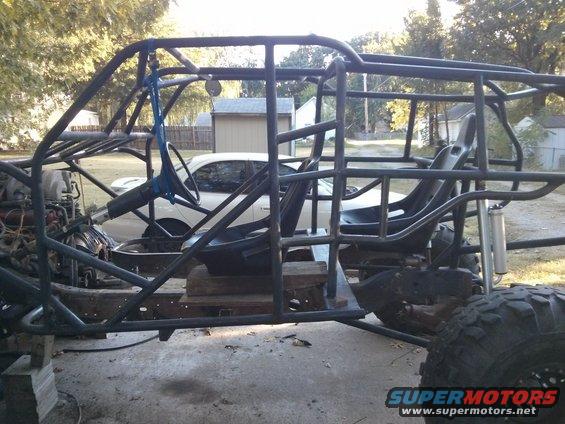 Roll cage frame of a custom buggy with seats and steering wheel installed. A white car is visible in the background