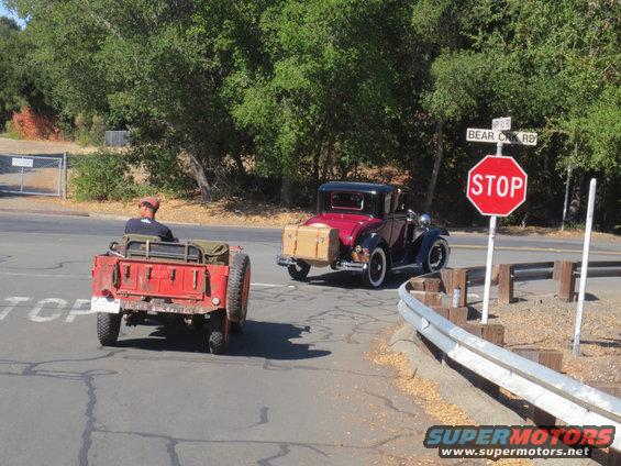 Red jeep-like vehicle with a trailer, seen from the rear, with a maroon coupe behind it