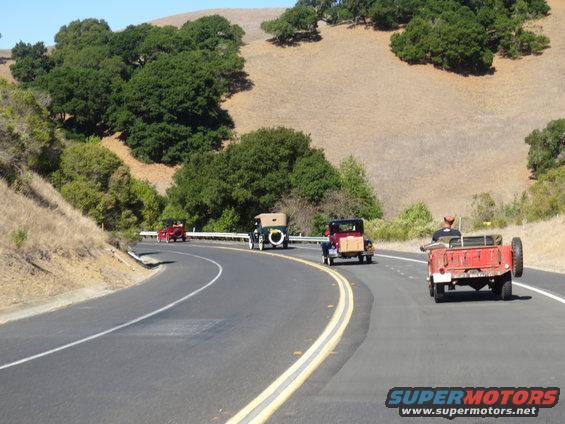 Four vintage cars driving on a rural road with hills and trees