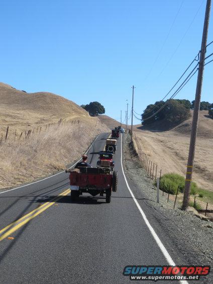 Red vintage truck with a driver, followed by other older vehicles on a rural road