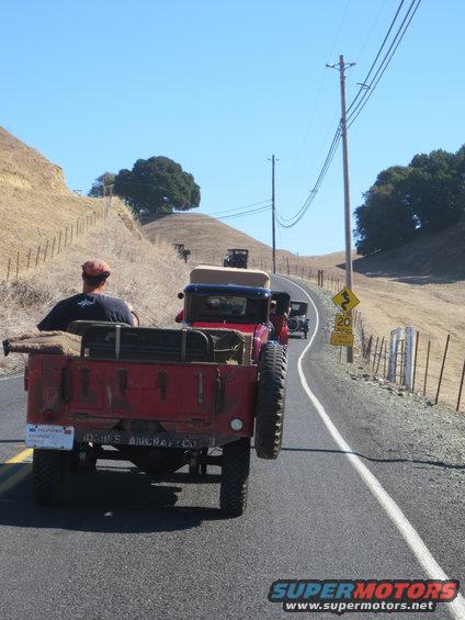 Red military-style truck with spare tire, driving on a rural road with other vintage vehicles
