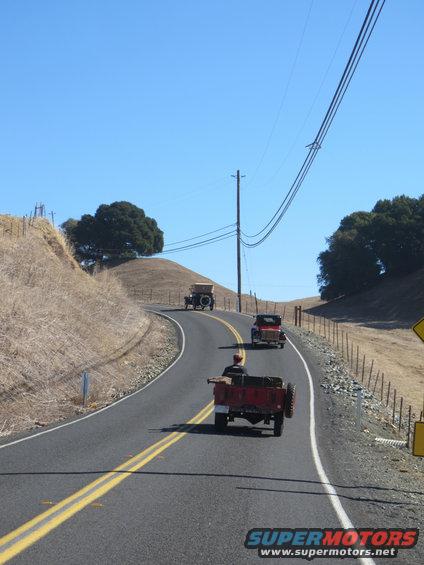 Three vintage vehicles, including a red trailer, drive on a two-lane road through a dry, hilly landscape