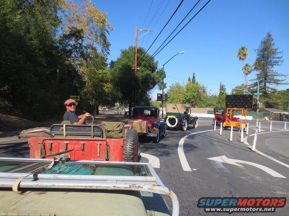 driver in flat cap in red jeep, other vintage cars behind on road