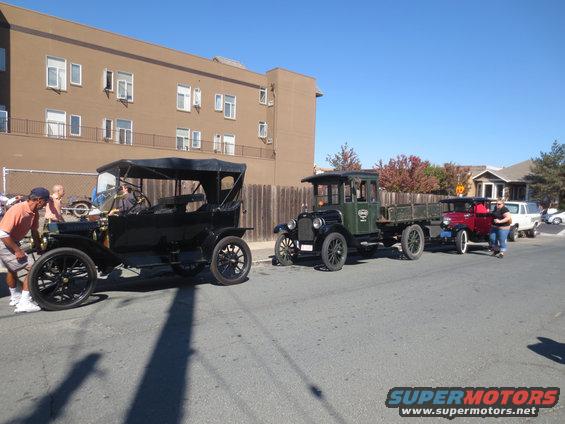 Three antique vehicles in a line on a street, a black touring car, a green truck, and a red truck