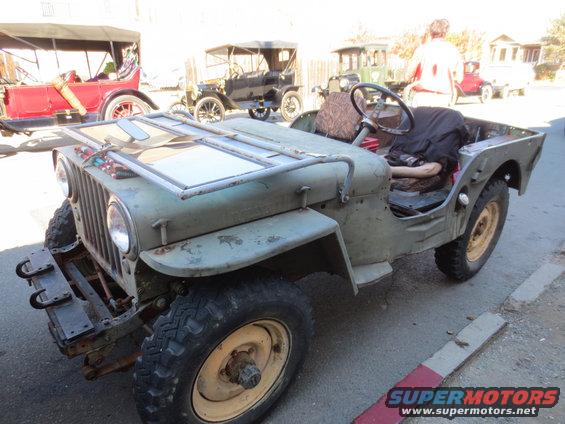 Olive green Jeep CJ-3A with a folded windshield and other vintage cars in the background
