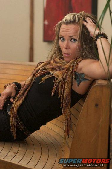Woman with blonde streaks in hair, wearing a scarf and black top, sitting on a wooden bench. Star tattoo visible on her arm