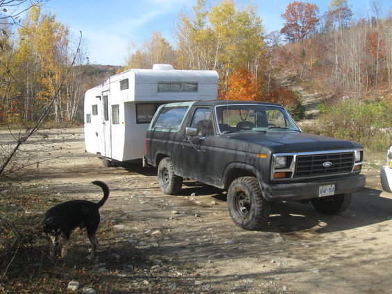 Black Ford Bronco towing a white travel trailer. License plate is visible. Dog stands in foreground