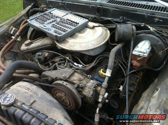Engine bay with a socket set on top of the air cleaner housing. Some hoses and belts are visible