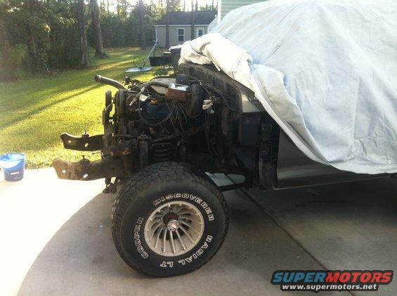 Vehicle chassis with engine exposed, partially covered by a tarp. Front wheel and tire visible