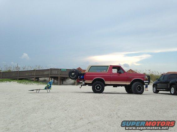Red 1994 Ford Bronco parked on a sandy beach with a wooden boardwalk in the background. Another dark SUV is parked to the right