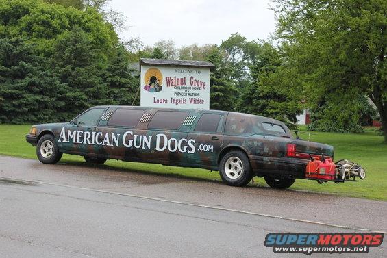 1992 Lincoln Town Car limousine with camouflage paint and 'AMERICAN GUN DOGS.COM' text, carrying gas cans and a small cart on the back