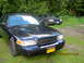 2003 Ford Crown Victoria - Two Ford Crown Victorias are parked on a grassy area with trees in the background. The front car is dark blue.
