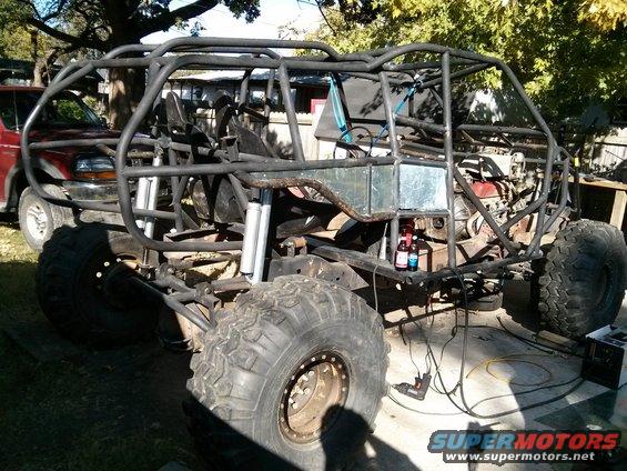 Custom off-road buggy frame with roll cage and large tires, red truck in background