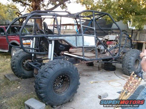 Custom buggy with roll cage, large tires, and exposed engine, parked next to a red pickup truck