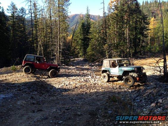 Two Jeep Wranglers, one red and one teal, on a rocky trail in a forest. The red Jeep is on the left, the teal Jeep is on the right