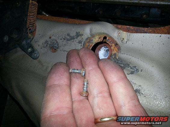 Hand holding rusty screws near a worn, tan seat bracket with a visible spring