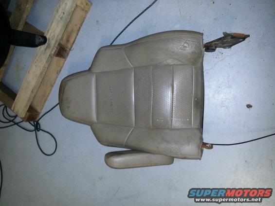 Tan leather car seat with perforated inserts and armrest, lying on a concrete floor next to a wooden pallet