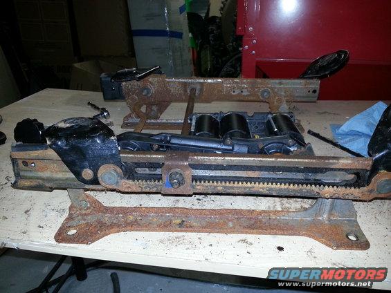 Two heavily rusted seat rail assemblies with black plastic components and metal gears on a workbench