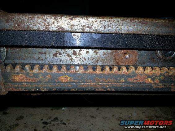 Close-up of a rusted metal seat adjustment mechanism with visible gear teeth and a black rubber strip