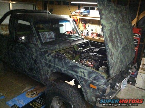 Camouflaged pickup truck with hood open, showing engine and interior. Visible text on a box below