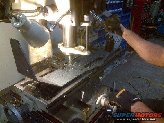 Milling machine with a metal plate being drilled, metal shavings on the base, and a person's gloved hand operating a crank
