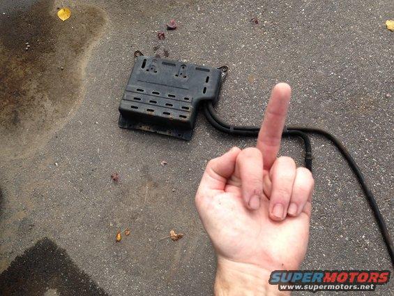 Black automotive part with hoses on asphalt next to a hand giving the middle finger. Water stain on left