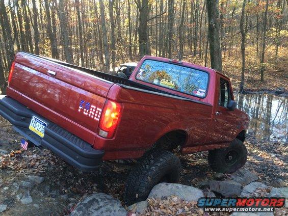 Red 1996 Ford F-150 pickup truck with lifted suspension driving over rocks in a wooded area