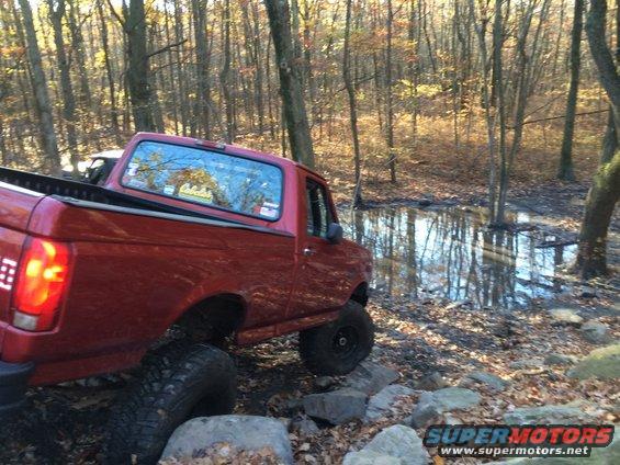 Red pickup truck with large tires navigating rocks and a muddy puddle in a wooded area
