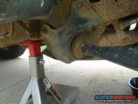 Undercarriage of a vehicle supported by a jack stand, showing rusty suspension components and a black oil drain pan
