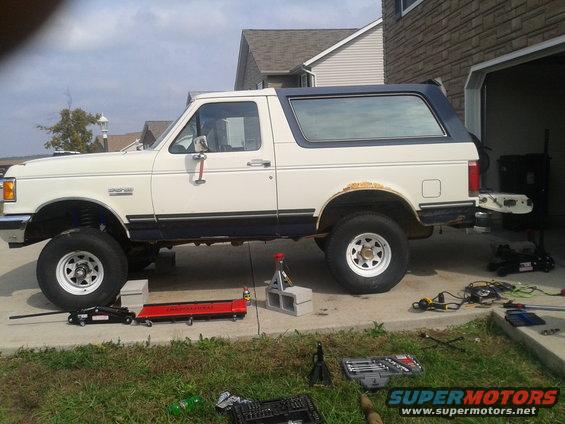 White Ford Bronco with blue trim, lifted, on jack stands, with rust on rear fender