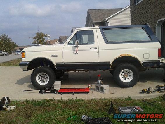 Side view of a white 1988 Ford Bronco with a blue top, lifted and on jack stands