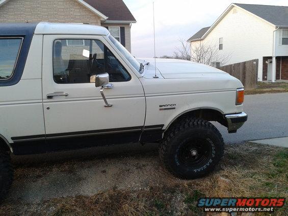 White Ford Bronco with dark trim and a blue top, parked on a dirt patch next to a house