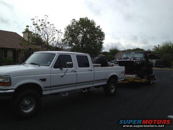 White crew cab pickup truck towing a black pickup truck on a trailer. The white truck appears to be a 1997 Ford F-350