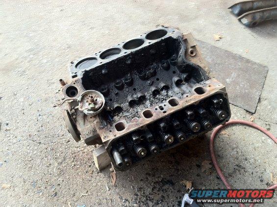 Engine block with visible valves and spark plug holes, resting on wooden blocks on a concrete surface