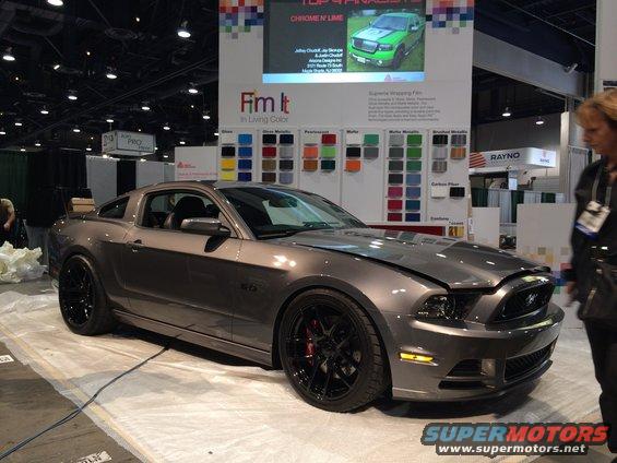 Gray Ford Mustang with black rims and red brake calipers. The hood is up, showing the engine bay. A display with color swatches is behind it