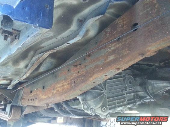 Underside view of a heavily rusted truck frame and transmission housing. Blue paint visible on upper frame rail