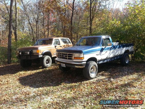 Two pickup trucks, a blue and white Ford and a brown Ford Bronco, parked outdoors in autumn. The blue truck is in the foreground