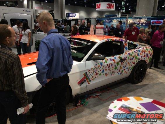 White 2013 Ford Mustang GT with orange hood and colorful square vinyl wrap, parked indoors at a show