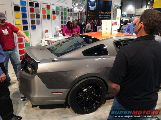 Gray sports coupe with black wheels and a spoiler, seen from the rear quarter angle at an auto show. People are looking at it