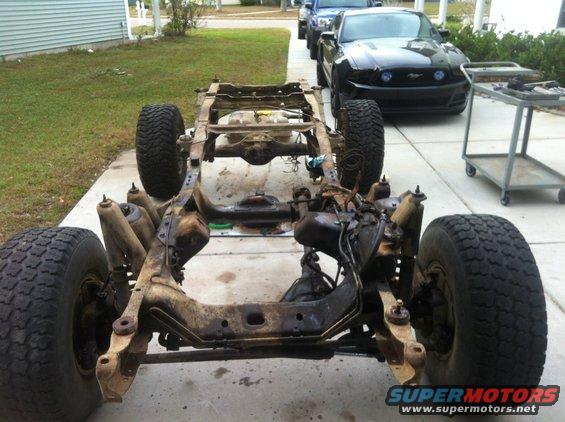 Vehicle frame and chassis with large tires. Black Ford Mustang in background