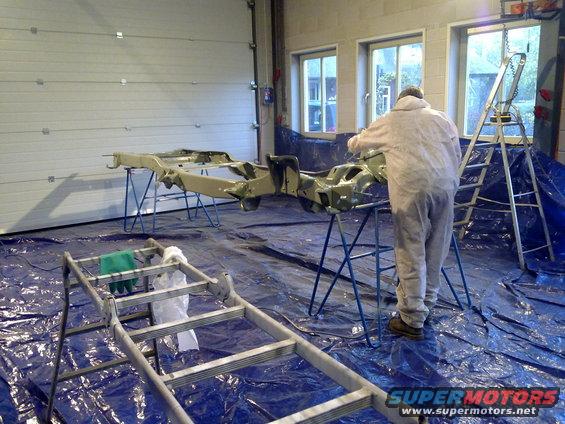 Vehicle frame on stands in garage, person in coveralls painting it