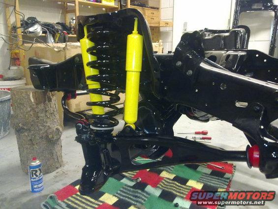 Close-up of a black vehicle frame with yellow shocks and black springs. Tools and a can of spray are nearby