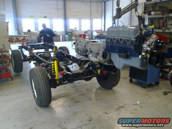 Vehicle chassis with yellow shocks and red suspension components, engine and transmission assembly being lowered by a crane