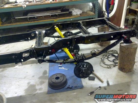 Black vehicle chassis on blocks, with yellow shock absorbers and brake drum. Tools scattered on floor
