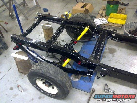 Black vehicle chassis with yellow shocks and red suspension components, on wooden blocks. Tires visible
