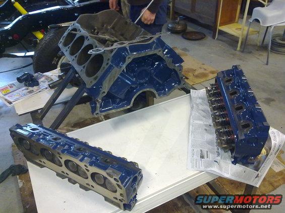 Engine block and two cylinder heads, all painted blue, sit on a table in a garage. A vehicle frame is visible in the background