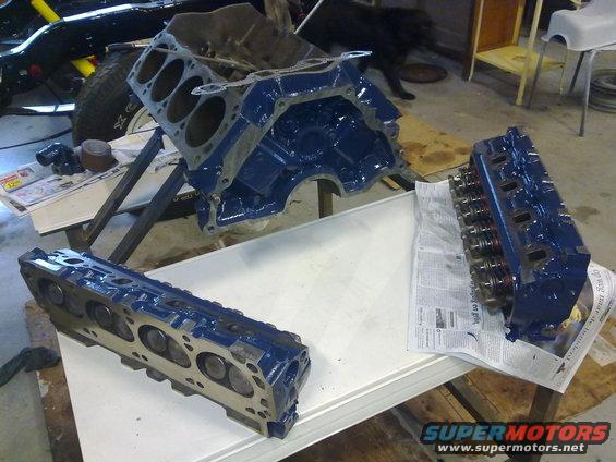Blue engine block and two cylinder heads on a table, with a partial vehicle in the background